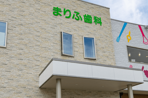 まりふ歯科医院の外観。明るいベージュのレンガ調外壁に緑色の看板文字が映える建物。
